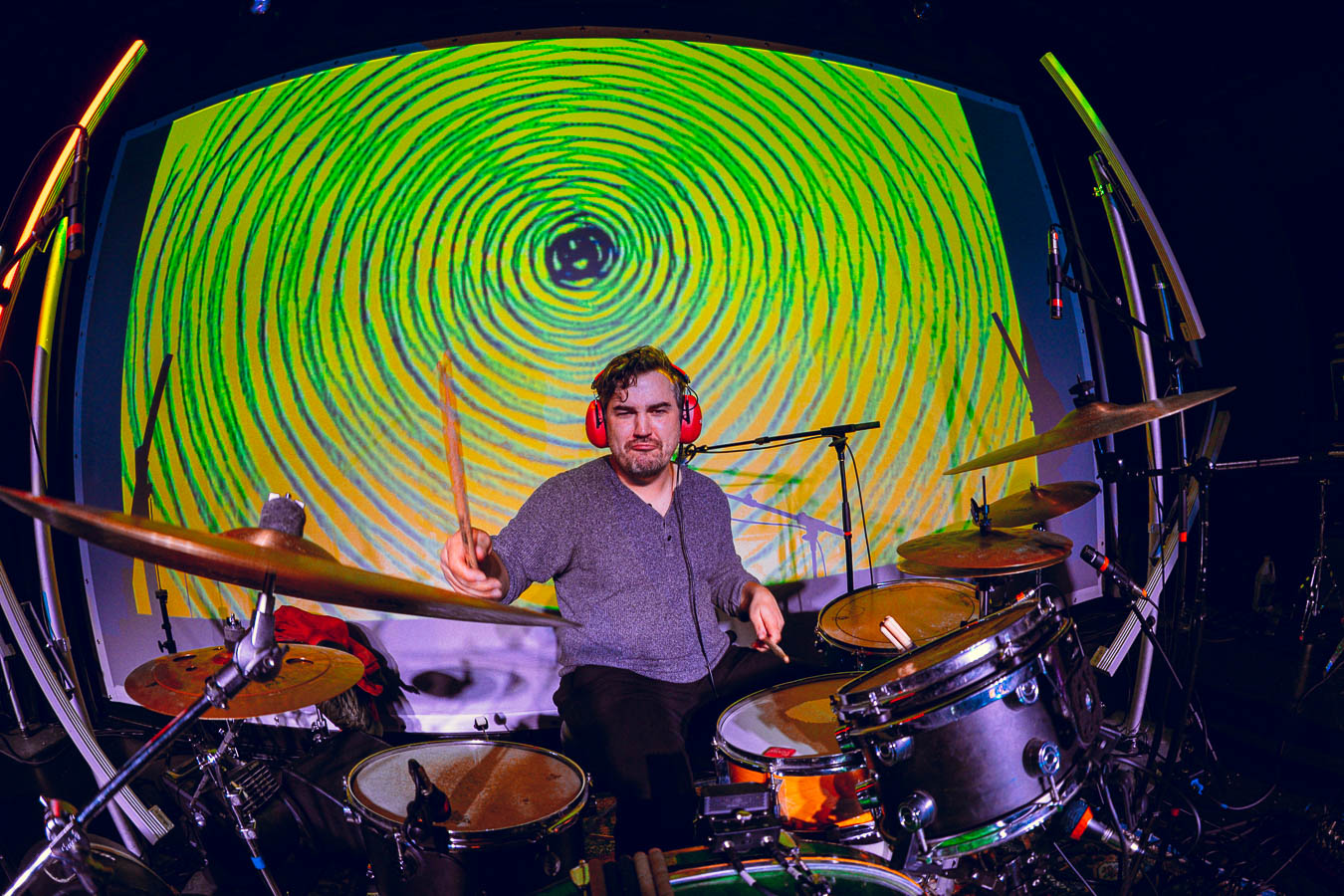 Max looks at the camera while he plays the drum set. The backdrop is a trippy green and yellow circular light projection.